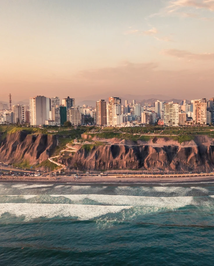A coastal cityscape with buildings along the shore, cliffs, and a beachfront with waves breaking in the foreground.
