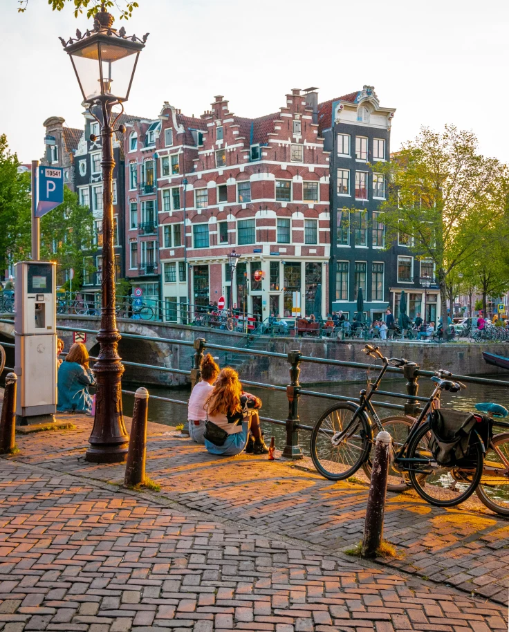 People sitting next to bikes and canal with buildings in the background during daytime
