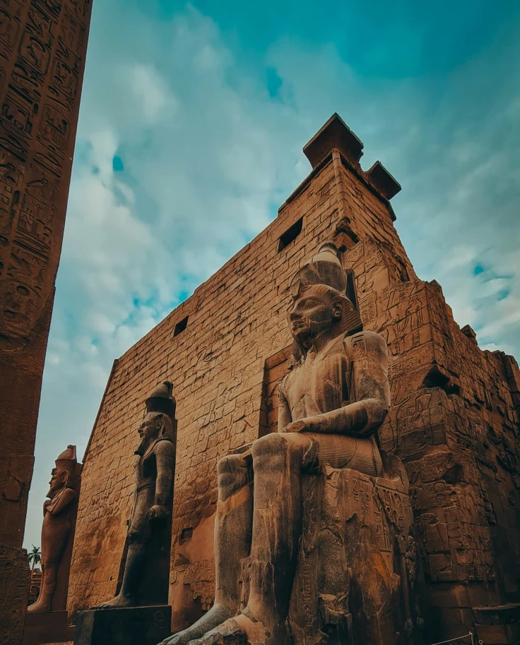 The image features grand ancient Egyptian statues seated before a temple, beneath a clear sky.