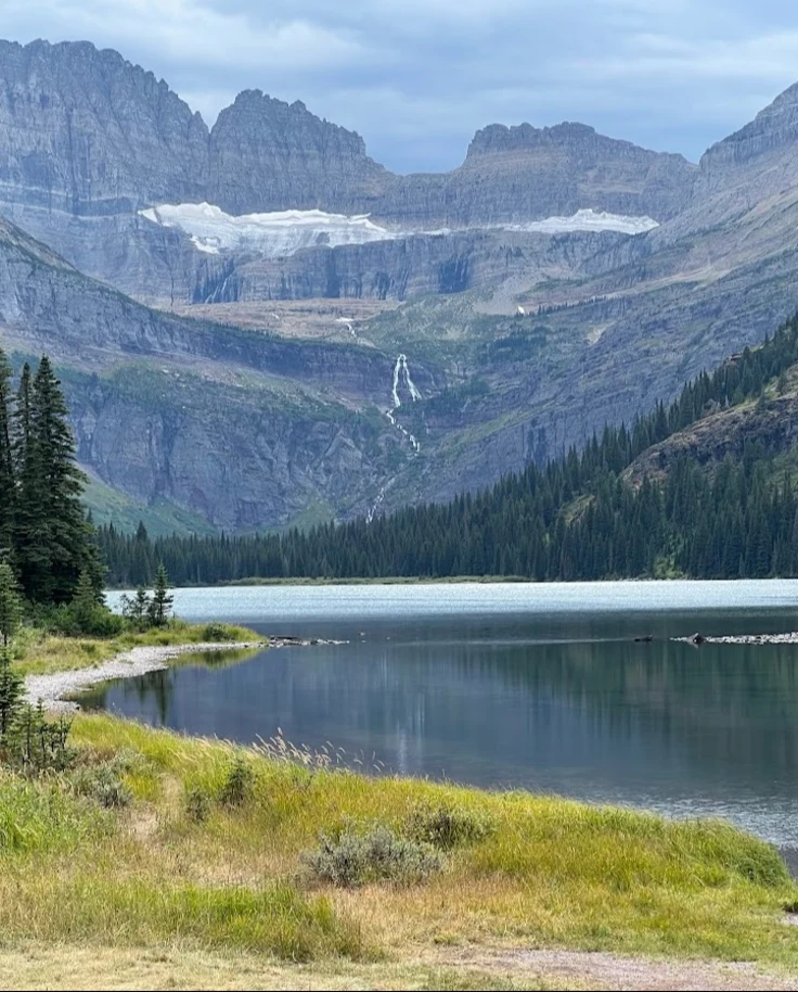 A serene view of a lake in Glacier National Park surrounded by mountains, green grass and trees.