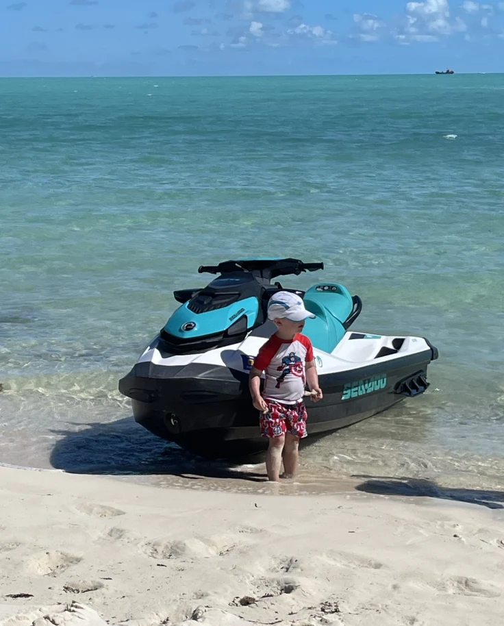 The image shows a person standing next to a blue and white jet ski on a sandy beach with the ocean in the background.