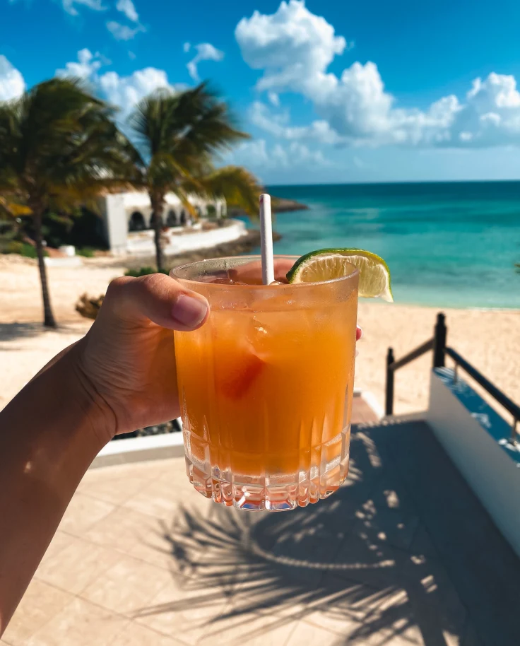 A hand presents a refreshing cocktail against a stunning beach backdrop, evoking a sense of tropical relaxation.