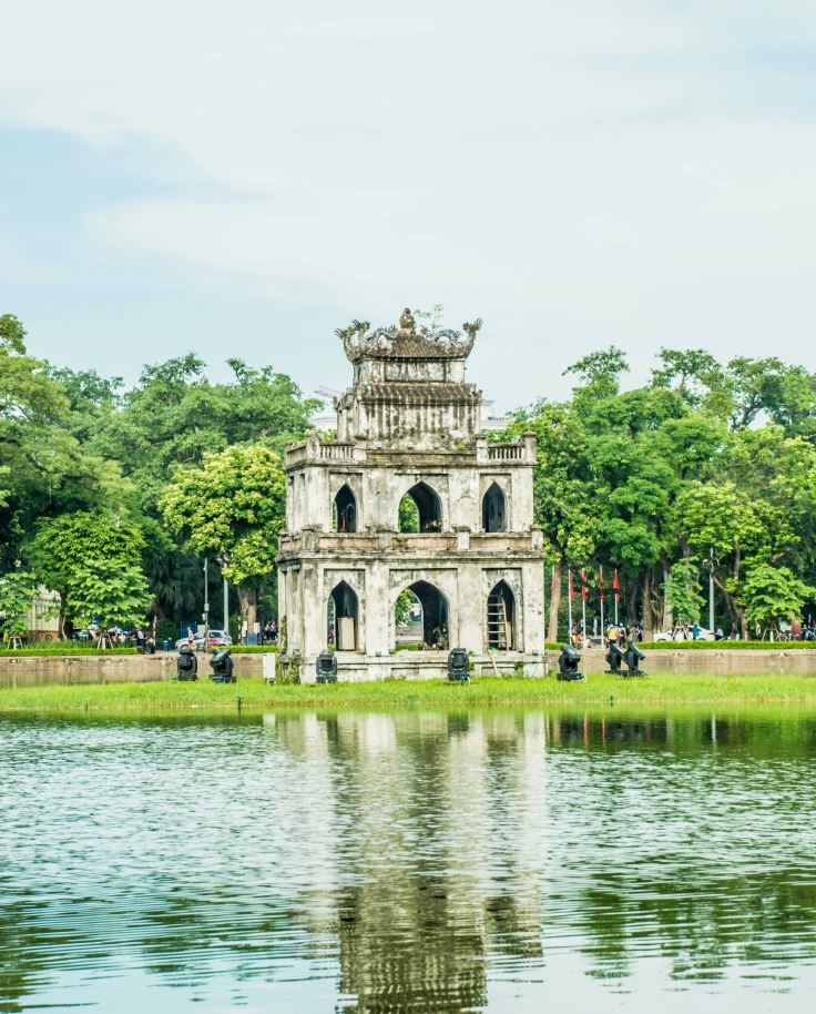 A three-tiered, ornate gate-like structure reflected in a body of water, surrounded by greenery and a clear sky.
