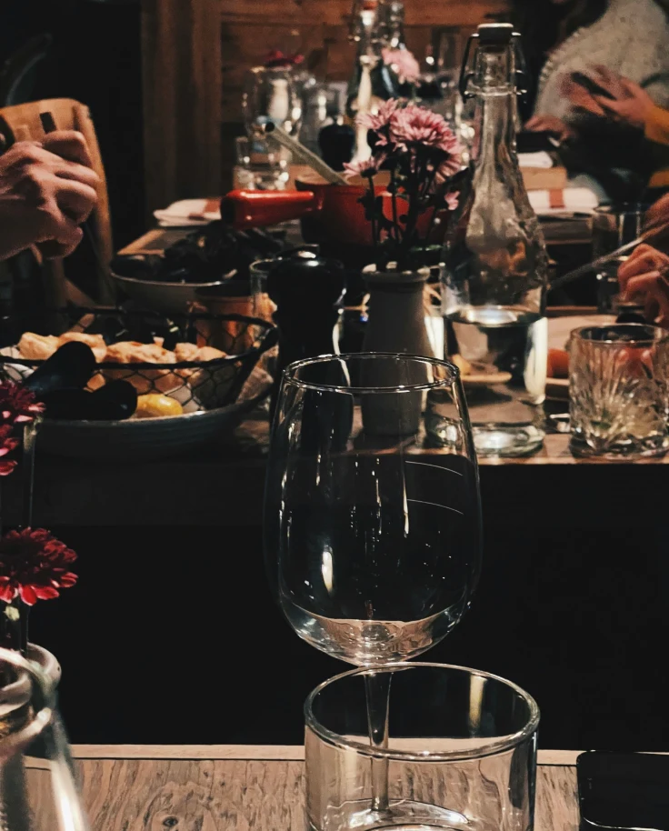 The image depicts a lively social dining scene with a table set with various glasses, bottles, and a vase with flowers.