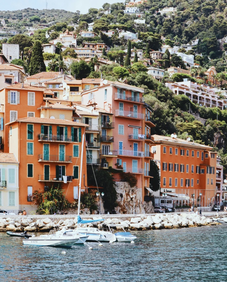 A view of the coastline with orange buildings and lush green landscape on the hillside with a boat sailing in view