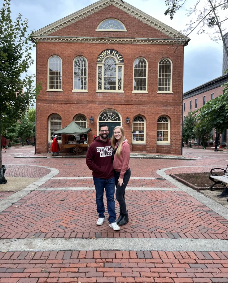 Two individuals pose in front of a brick building with arched windows and a circular window at the top center.