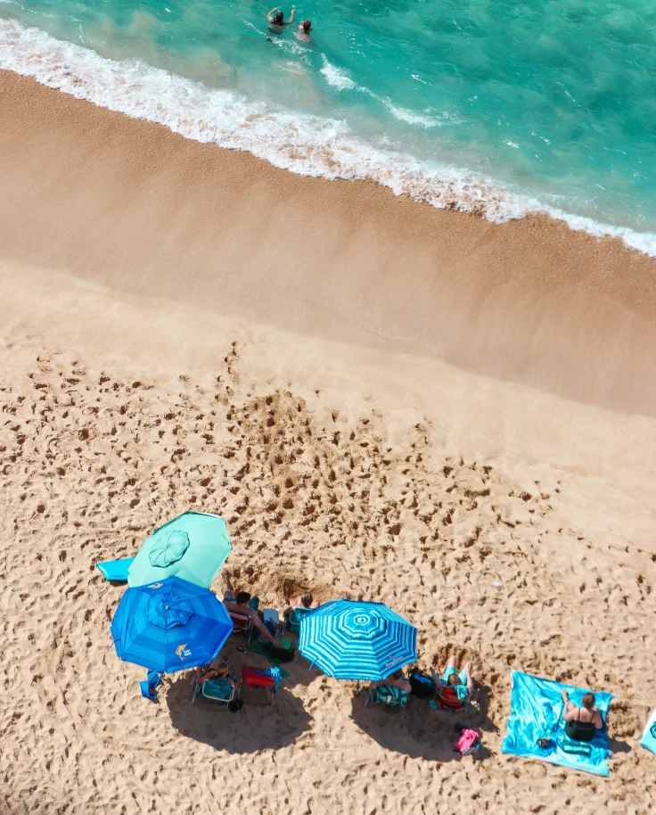 An aerial view of the beach with some of the people lying on the sand under beach umbrellas, while others swimming in the water.