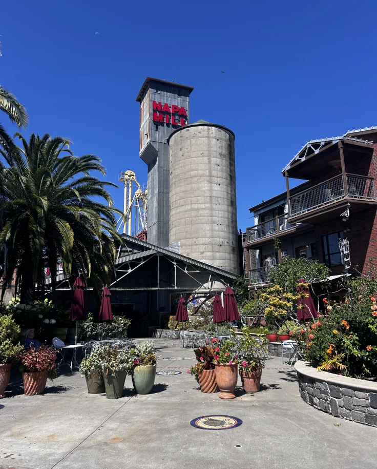 Napa Mill — a historic mixed-use complex featuring boutique shops, dining, and luxury accommodations along the Napa River — with a tall metal tower in the center of a concrete courtyard with trees.