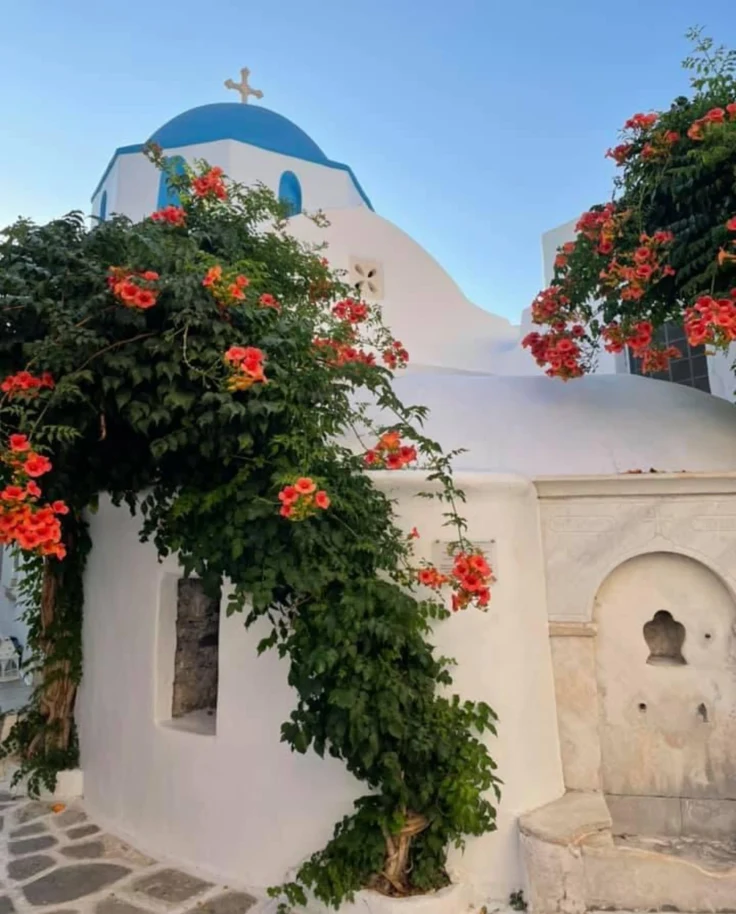 The image portrays a white building with a blue dome, adorned with red flowers and greenery, reflecting classic Greek architectural beauty.