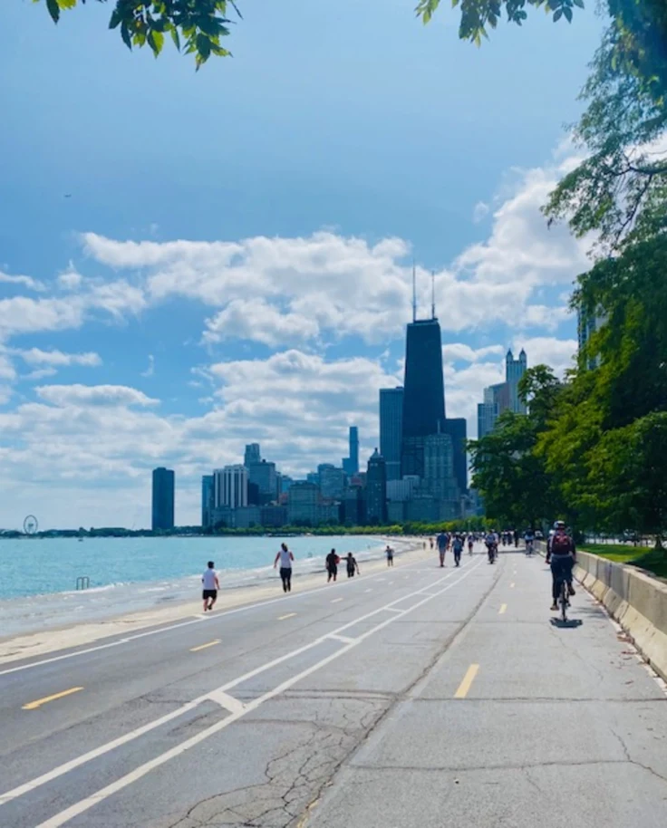 The image offers a picturesque view of a cityscape from a lakeside road, with pedestrians and cyclists enjoying the clear blue sky with scattered clouds.