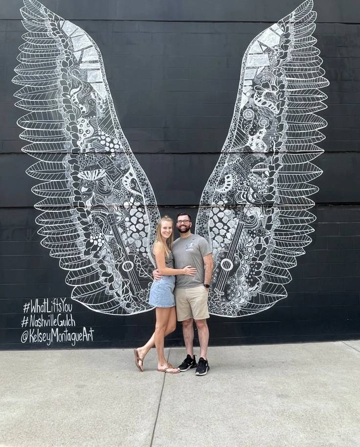 A couple posing for a photo in front of a wall art mural of angel wings