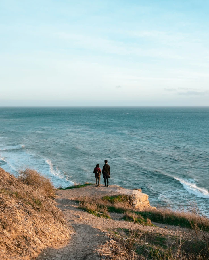 two people standing on a cliffside next to the ocean during the daytime