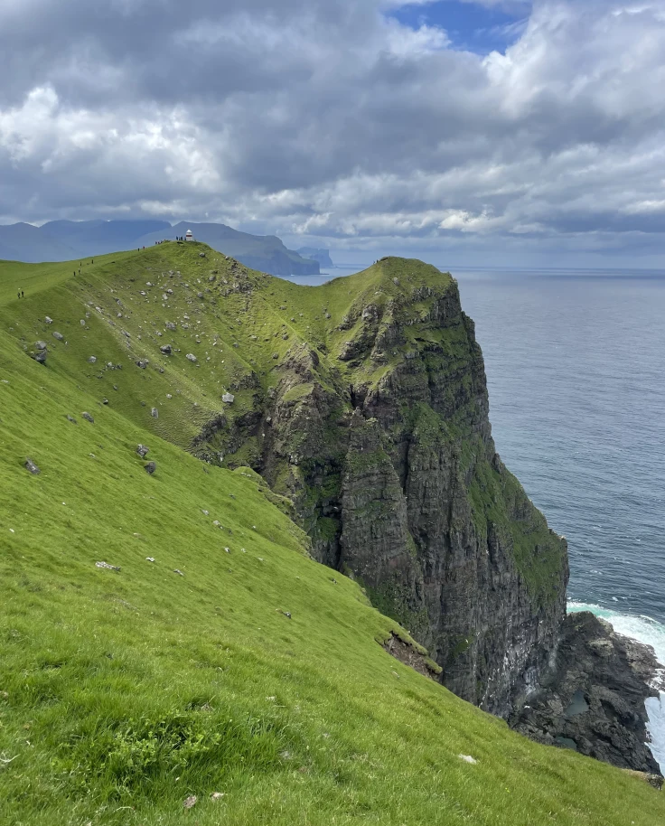 A view of grassy and rocky cliffs with the sea and a cloudy blue sky in the background.