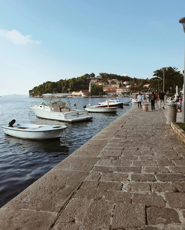 A picturesque promenade by the water with moored boats and a hillside town in the background.