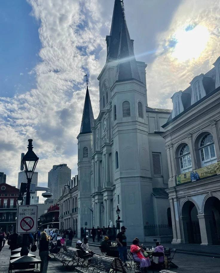 A church with tall steeples and benches in front of it with the sun peeking through clouds behind it.