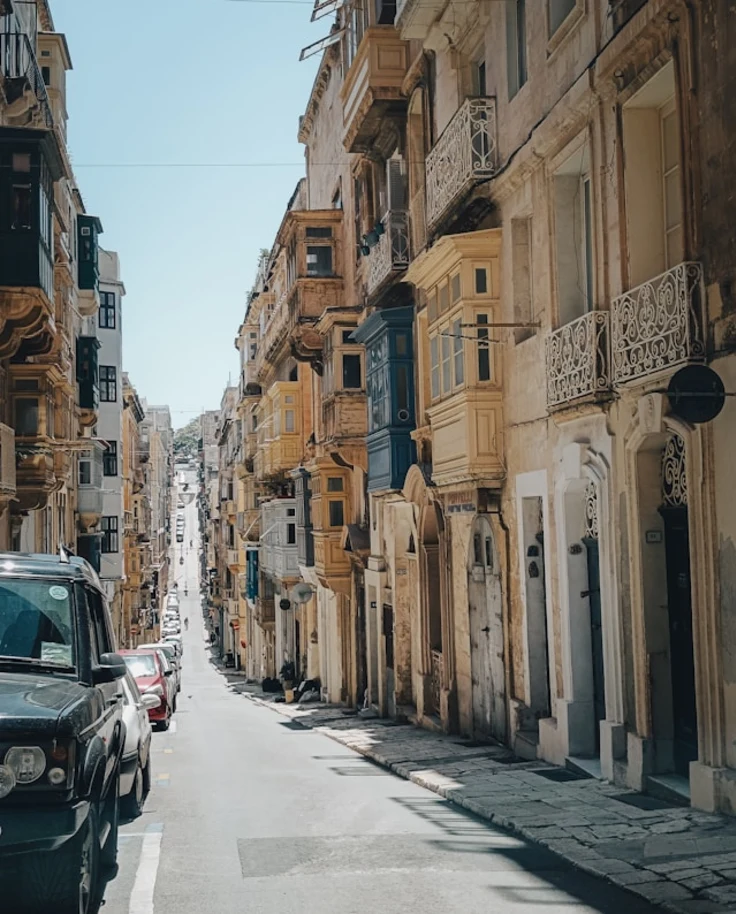 An image of a European city street with various stone balconies and detailing. There are cars parked along the left side of the street.