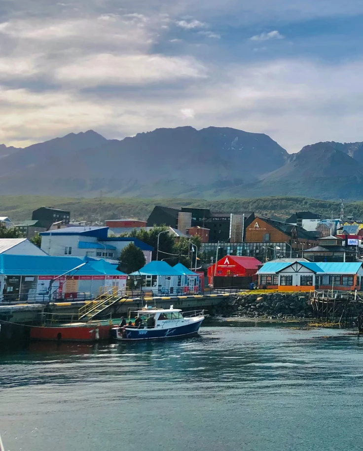 A blue lake with pink, blue and yellow houses and boats with green and brown mountains in the distance in Ushuaia on a Argentina vacation.