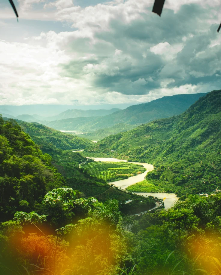 Forested mountains surround a lush green valley with a winding river under a cloudy sky.