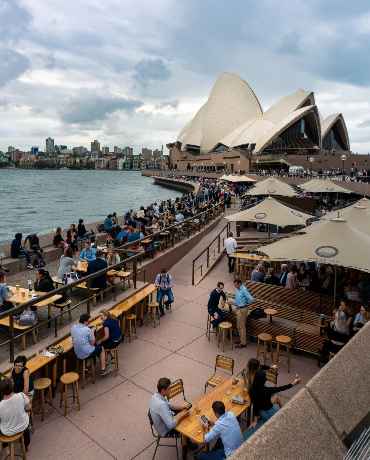 The image depicts an outdoor dining scene with guests enjoying a meal against the backdrop of the Sydney Opera House under a cloudy sky.