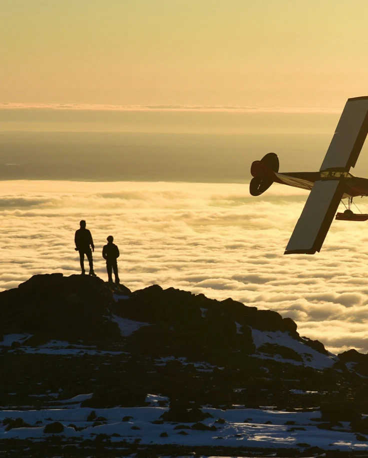 Two people stand on a rocky outcrop above a sea of clouds, with an airplane flying nearby during a stunning sunset or sunrise.