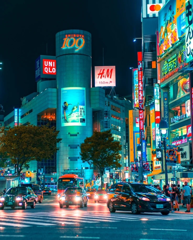 A view of city buildings lit up at night in Japan surrounded by cars and tourists.