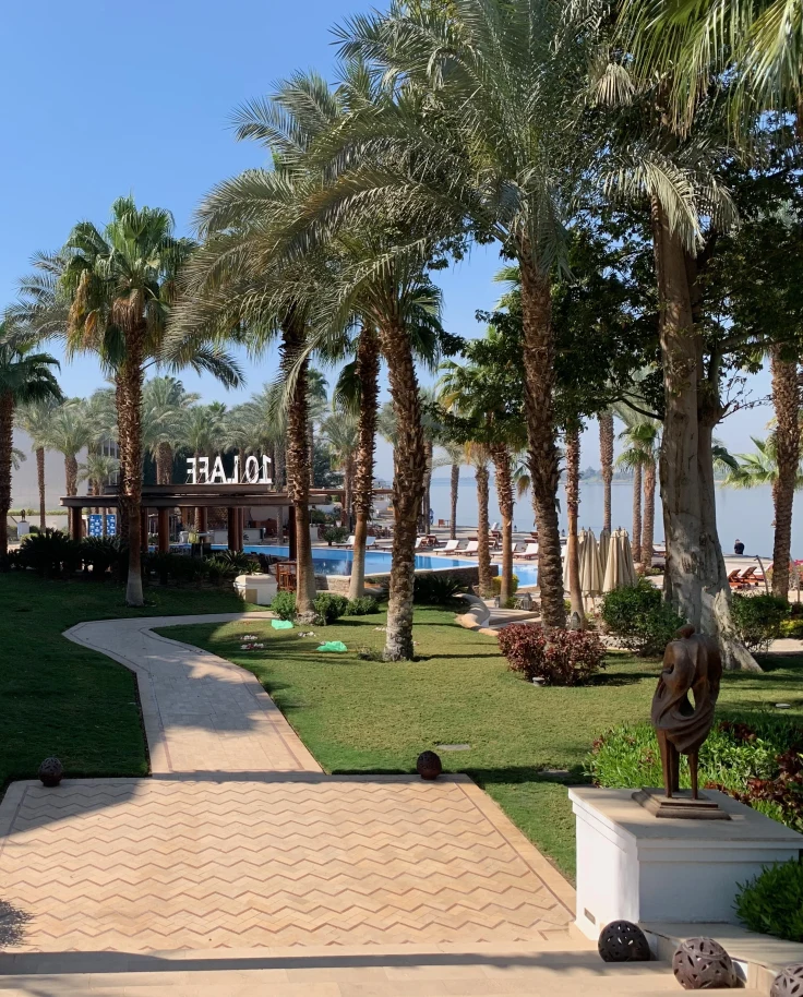 The image presents a landscaped pathway lined with palm trees and sculptures, leading to a building.