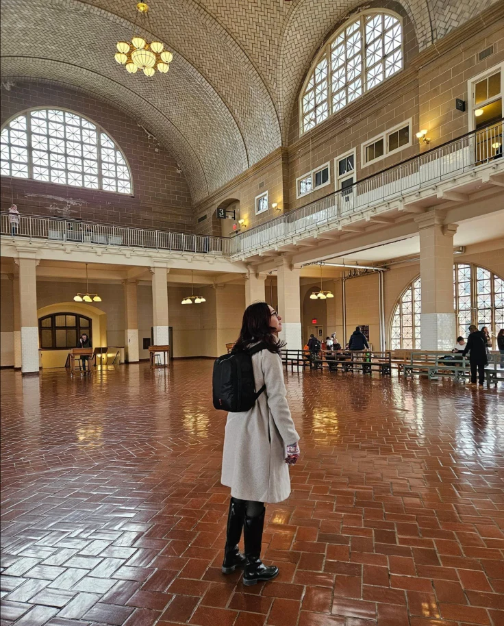 A woman standing in an open plaza that looks like a train station atrium with a gray coat and black backpack looking up at the ceiling.