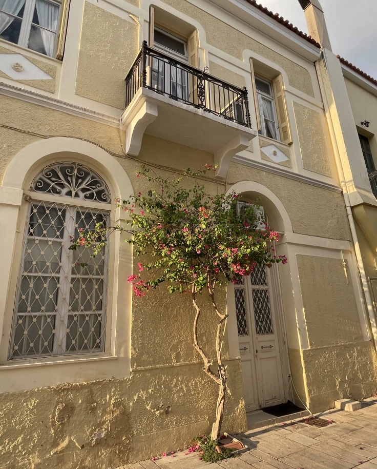 The image shows the exterior of a beige building with decorative windows, a small balcony and flowering plants.