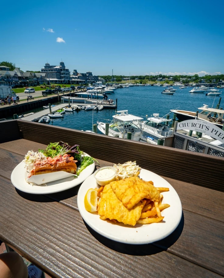 A view of two plates of food on a ledge overlooking a marina with boats docked on a sunny day.