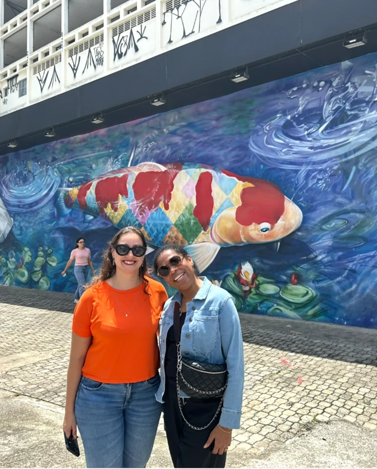 The image captures two people posing before a colorful mural depicting a large koi fish surrounded by marine life.