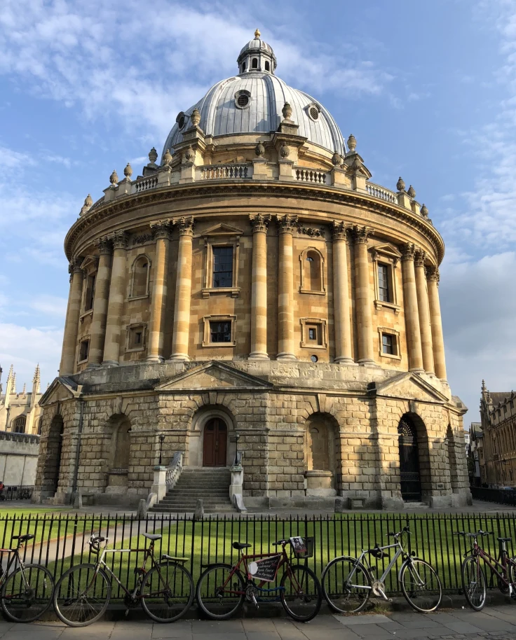 A view of a large dome building with ornate architexture and an iron gate surrounding it, known as the Radcliffe Camera, during the day.