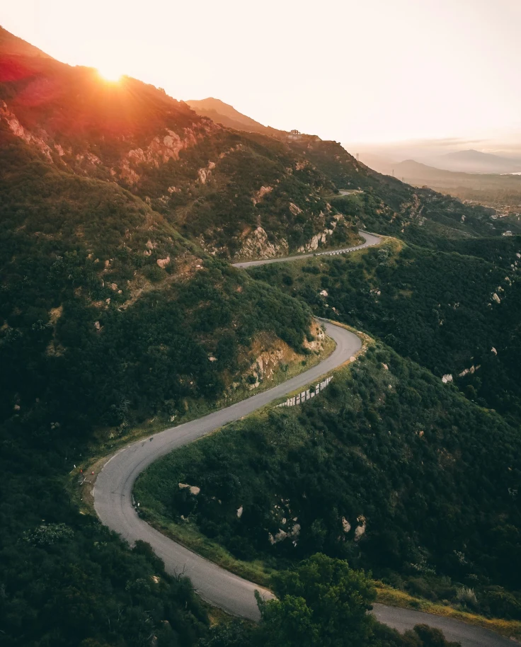 An aerial view of a winding road through green mountains during the sunrise.