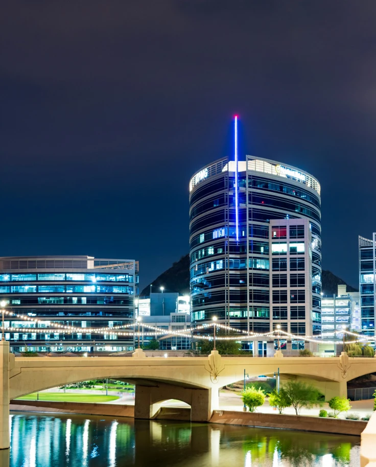 A city skyscraper lit up at night with a large stone bridge over a river next to it.