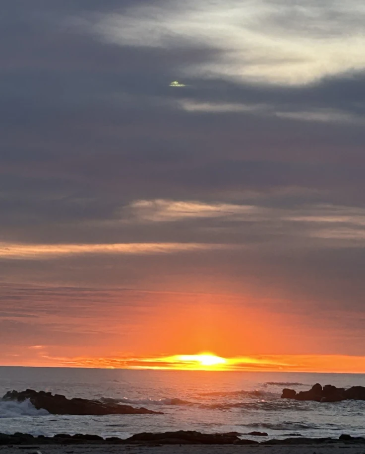 A sunset over the ocean with the sun partially obscured by the horizon and clouds, creates a vibrant display of colors in the sky.