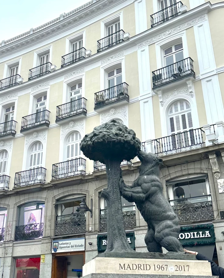 A poignant depiction of a bear reaching for a strawberry tree, set before a classic building with commercial undertones in Madrid.