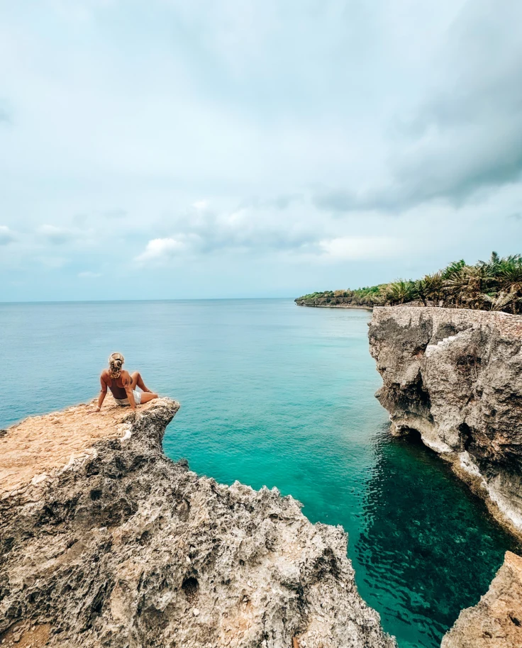 A picture of a person sitting on a rocky cliff near the water during the daytime.