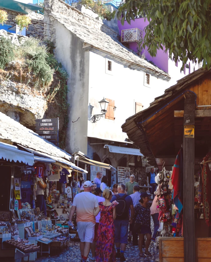 A picture of people walking down a cobblestone street with various markets, stands and shops surrounding them on the left and right sides. There is a white building with a slanted roof in the distance, and trees near it.