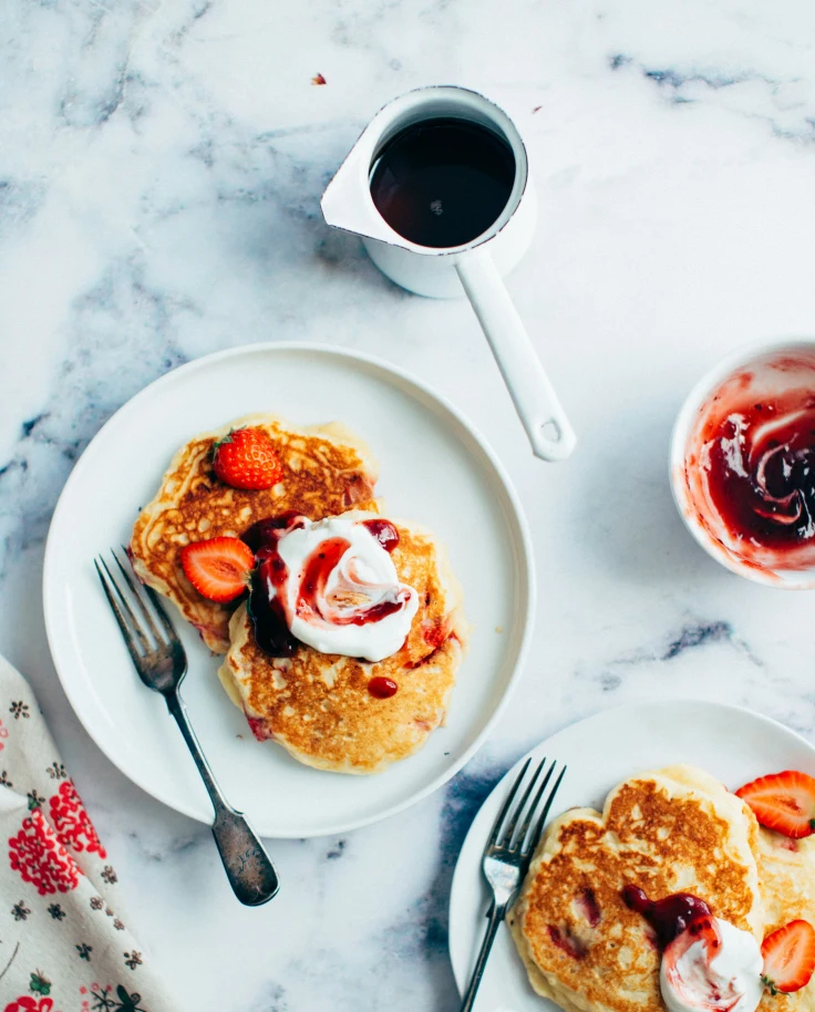 Plates of pancakes topped with whipped cream and berries on a white marble table.