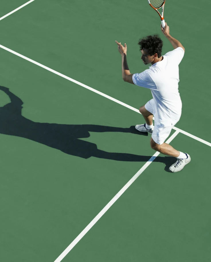 A tennis player in white attire mid-action on a green tennis court on a sunny day.