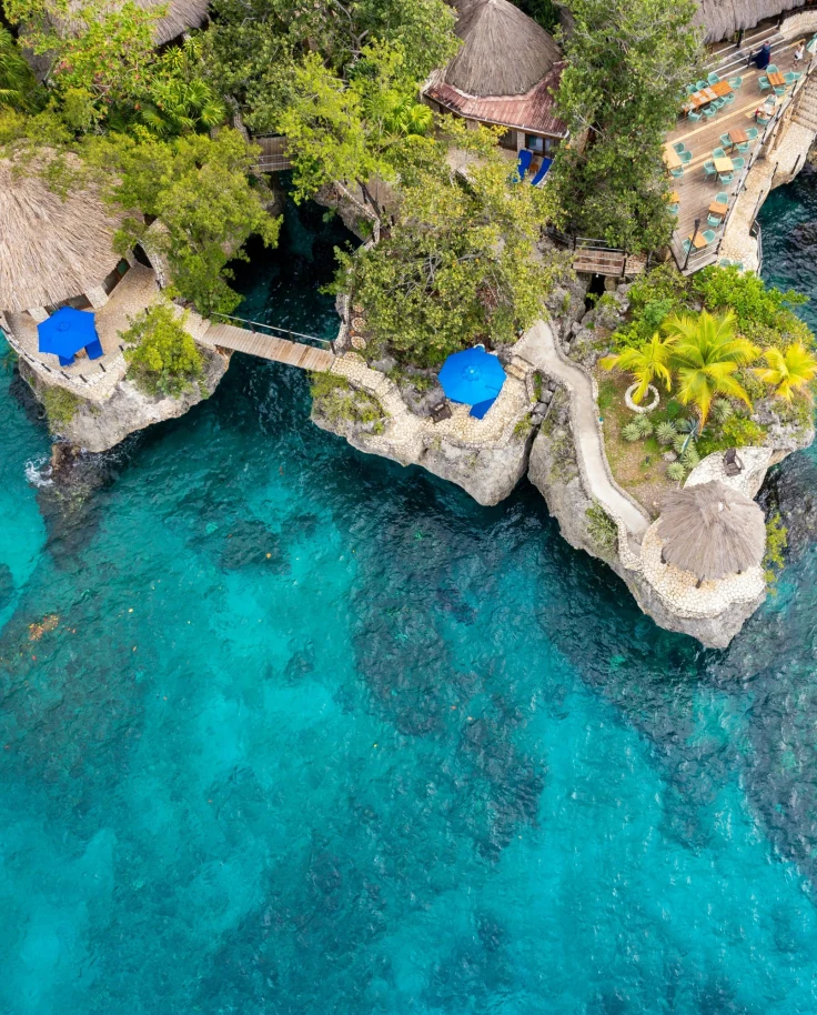 The image is an aerial view of a coastal resort with thatched-roof structures and blue umbrellas, surrounded by clear turquoise waters.