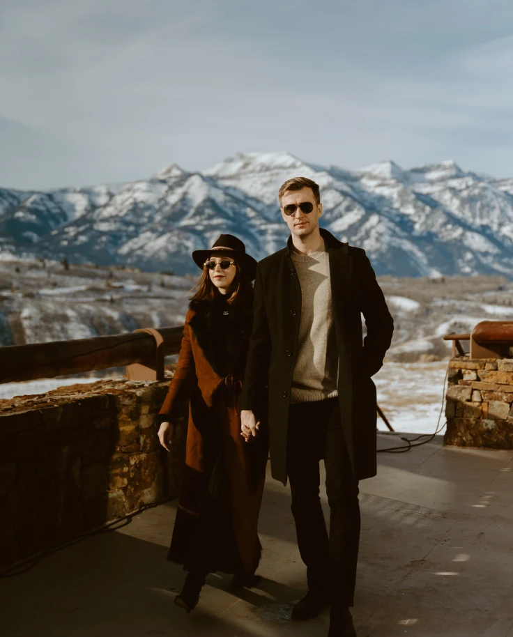 A couple dressed in swanky winter clothes, standing on an open balcony, posing in front of snow-covered mountains.