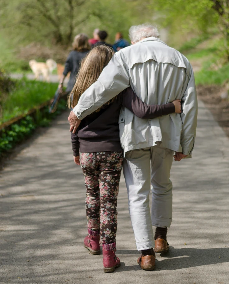 Two companions walk arm in arm along a path surrounded by lush greenery.
