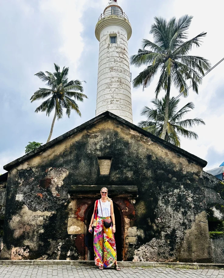 A picture of a person standing in front of an old lighthouse with palm trees under a cloudy sky.
