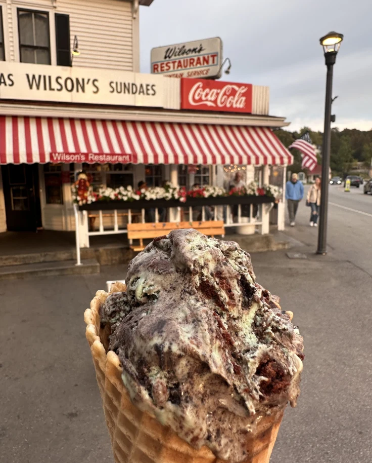 A close-up view of an ice-cream cone in front of a store with red and white awning.
