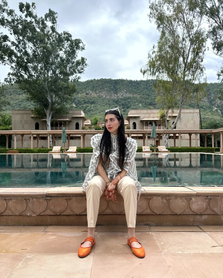 A woman wearing orange shoes, white shorts and a patterned blue top seated on a stone ledge in front of a swimming pool with trees and a mountain in the background.