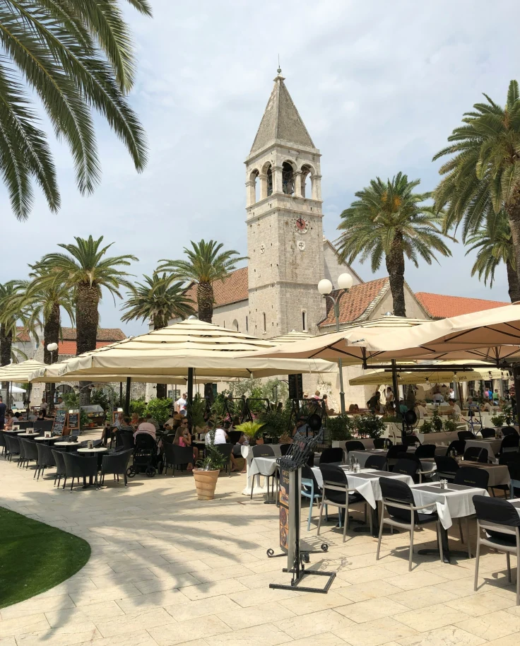 A bustling outdoor café scene with diners under umbrellas, flanked by palm trees and a historic bell tower in the background.