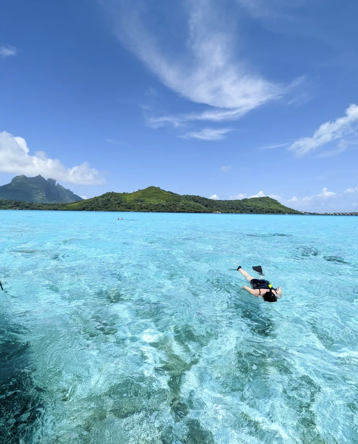 A person snorkeling on the crystal clear blue water with a green mountain in the background.