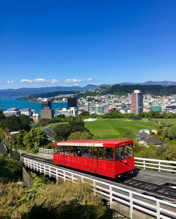 Red bus driving on a bridge with city buildings and sea in the background.