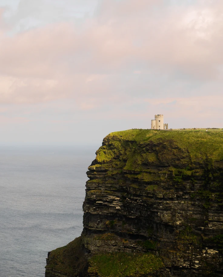 Green cliff with white building next to body of water with pink clouds in the sky during daytime