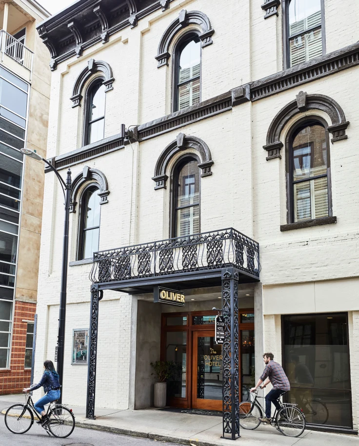The entrance of a three story white building with black trim called the Oliver hotel and two people riding bikes during the day.